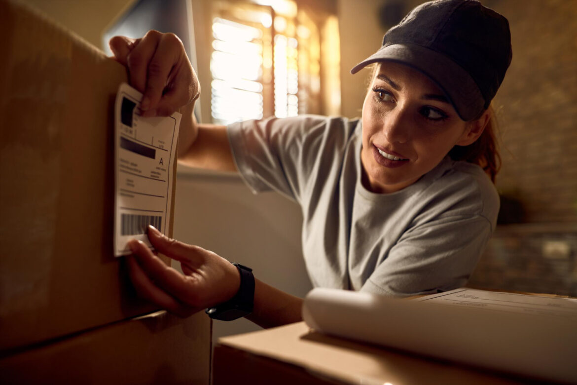 joven-repartidora-preparando-paquetes-envio-adjuntando-etiqueta-datos-caja-carton-oficina devoluciones